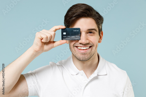 Young happy smiling cheerful fun caucasian man 20s wearing white casual basic t-shirt covering eye with credit bank card isolated on pastel blue background studio portrait People lifestyle concept