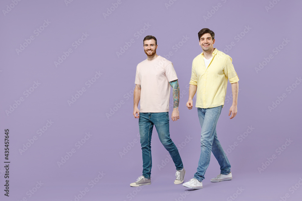 Full length side view two young happy men 20s friends together in casual t-shirt walking going strolling lokk camera isolated on purple background studio portrait People friendship lifestyle concept Full length side view two young happy men 20s friends together in casual t-shirt walking going strolling lokk camera isolated on purple background studio portrait People friendship lifestyle concept