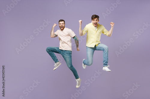 Full length two young overjoyed cool men friends together in casual tshirt do winner gesture clench fist jump high celebrating scream yes isolated on purple background studio People lifestyle concept