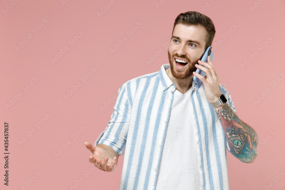 Young happy smiling caucasian unshaven man in blue striped shirt talking by mobile cell phone conducting pleasant conversation spread hands isolated on pastel pink background People lifestyle concept Young happy smiling caucasian unshaven man in blue striped shirt talking by mobile cell phone conducting pleasant conversation spread hands isolated on pastel pink background People lifestyle concept
