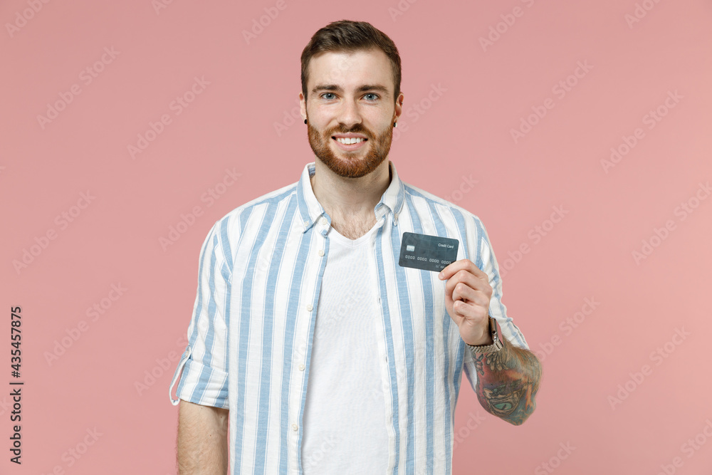 Young smiling fun rich successful happy caucasian unshaven man in blue striped shirt hold credit bank card looking camera isolated on pastel pink background studio portrait. People lifestyle concept. Young smiling fun rich successful happy caucasian unshaven man in blue striped shirt hold credit bank card looking camera isolated on pastel pink background studio portrait. People lifestyle concept.