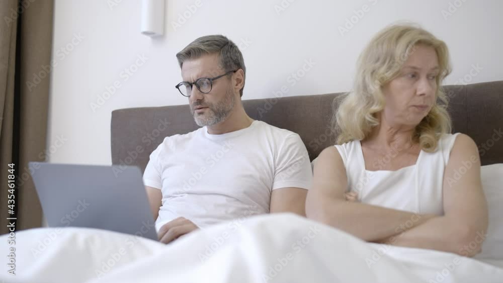 Blond wife lying in bed and trying to talk to busy husband working on laptop Blond wife lying in bed and trying to talk to busy husband working on laptop