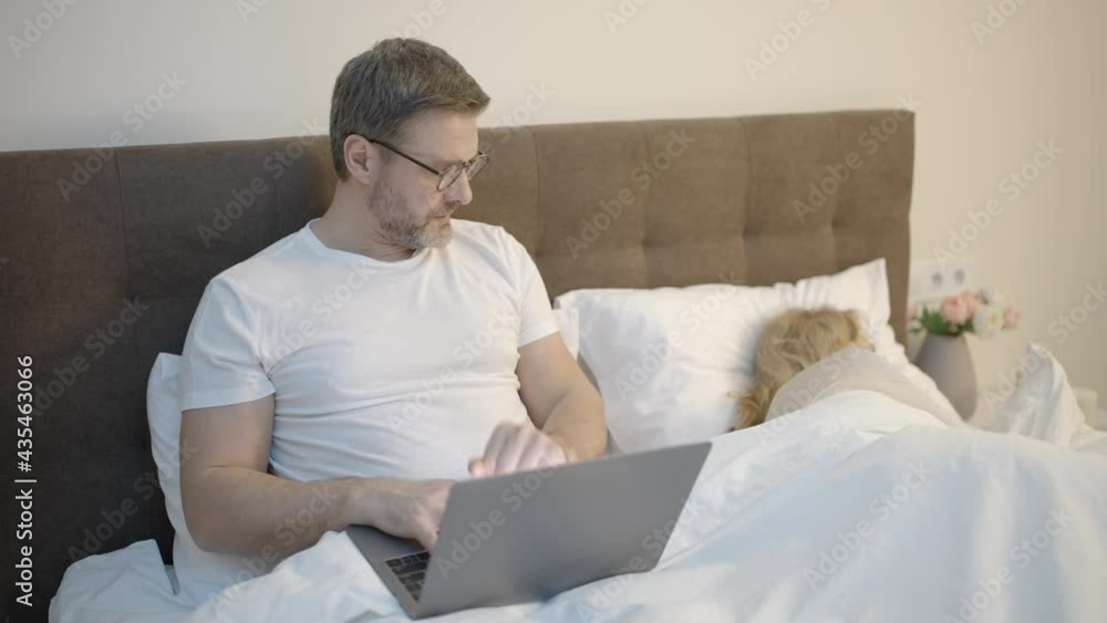 Man staying up late in bed near sleeping wife, typing on laptop, workaholic Man staying up late in bed near sleeping wife, typing on laptop, workaholic