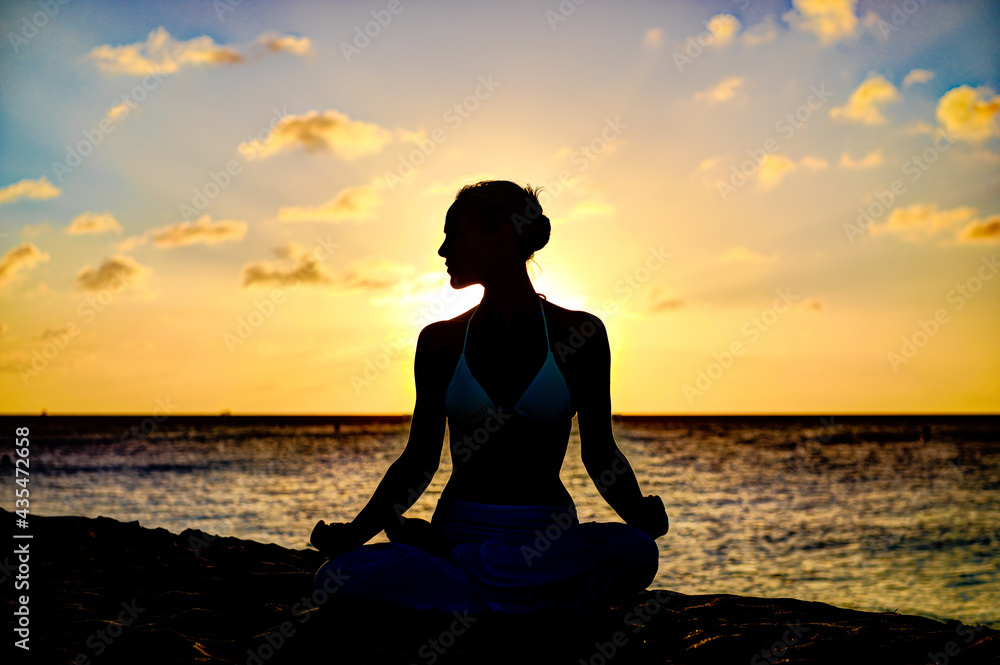 Woman meditating, doing yoga, at the beach, sitting by the seashore, dressed in a white outfit at sunset Woman meditating, doing yoga, at the beach, sitting by the seashore, dressed in a white outfit at sunset