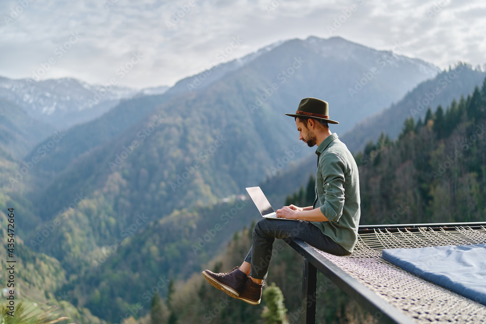 Young man freelancer traveler wearing hat anywhere working online using laptop and enjoying mountains view - obrazy, fototapety, plakaty 