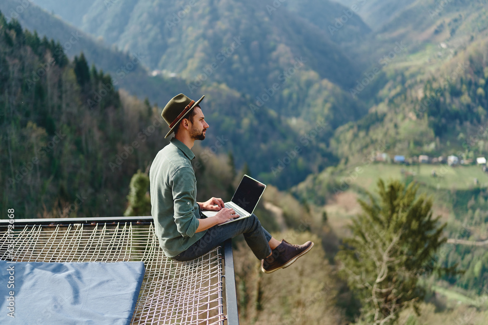 Young man freelancer traveler wearing hat anywhere working online using laptop and enjoying mountains view - obrazy, fototapety, plakaty 