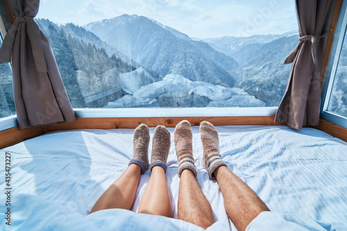 Lover couple in knitted warm socks lying on a soft cozy bedroom with snowy mountains view in a winter time