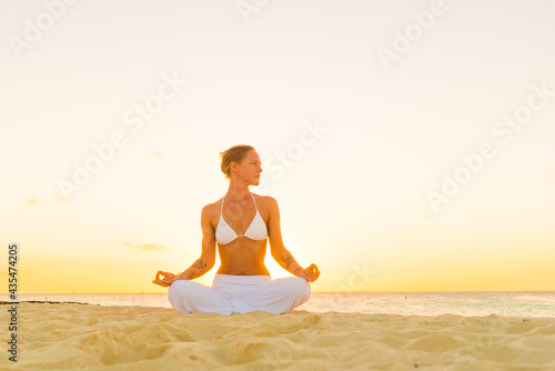 Woman meditating, doing yoga, at the beach, sitting by the seashore, dressed in a white outfit at sunset © TRAVEL EASY