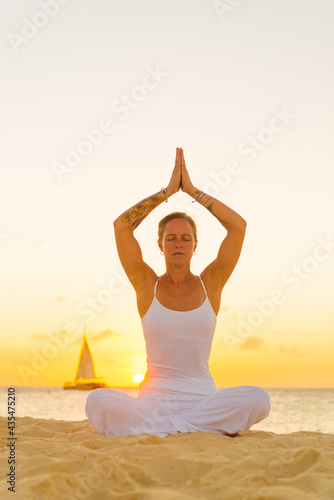 Woman meditating  doing yoga  at the beach  sitting by the seashore  dressed in a white outfit at sunset