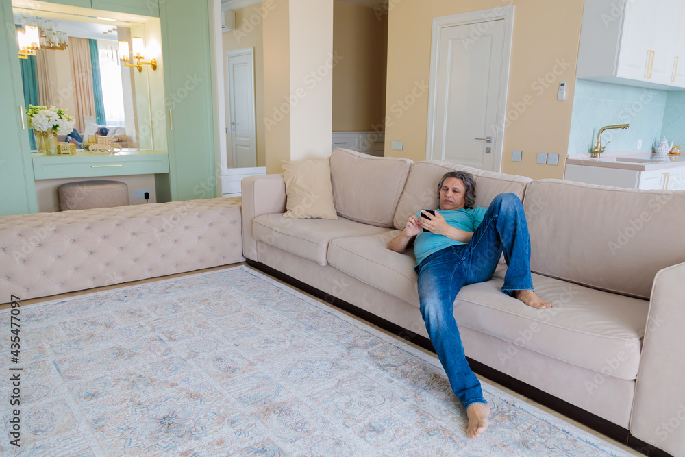 An adult man browses social networks in a mobile phone while lying on a sofa in an apartment. An adult man browses social networks in a mobile phone while lying on a sofa in an apartment.