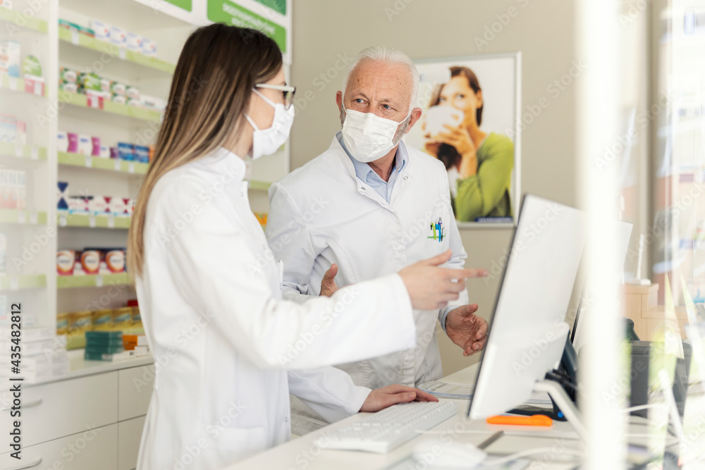 Fototapeta premium Conversation between two pharmacists and a corona virus. An elderly pharmacist and an adult pharmacist stand behind the counter at the pharmacy and sell medicines. They wear uniforms and face masks