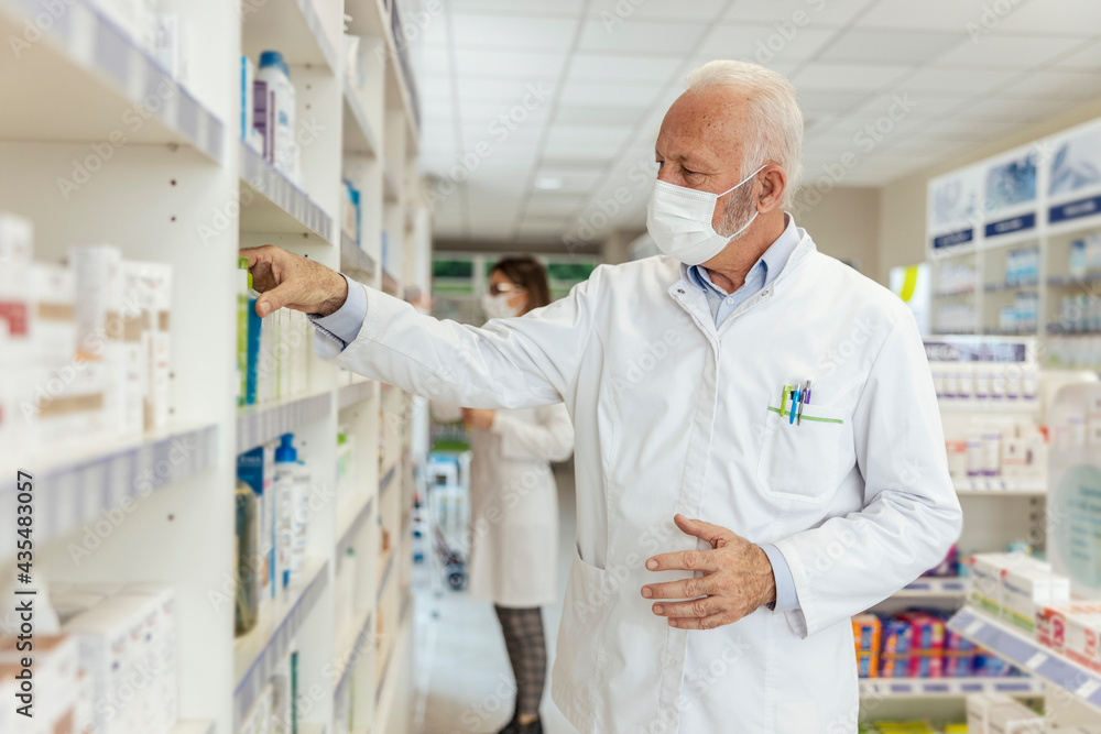 Buying and selling medicines, doctor's advice and help to find medicines on the shelf. Close up of a man with gray hair in a white uniform and a protective face mask sorting drugs at the pharmacy Buying and selling medicines, doctor's advice and help to find medicines on the shelf. Close up of a man with gray hair in a white uniform and a protective face mask sorting drugs at the pharmacy