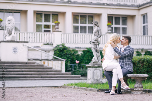 A tender beautiful newlywed couple sits on a bench against the backdrop of greenery and a castle.