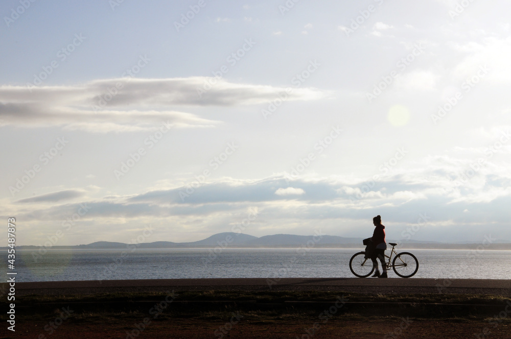 A woman walking with her bicycle on the side of the path. In the background you can see the sea and some mountains A woman walking with her bicycle on the side of the path. In the background you can see the sea and some mountains