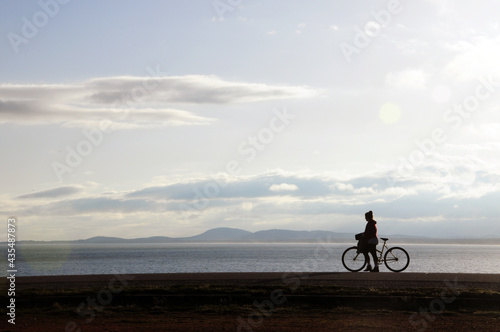 A woman walking with her bicycle on the side of the path. In the background you can see the sea and some mountains