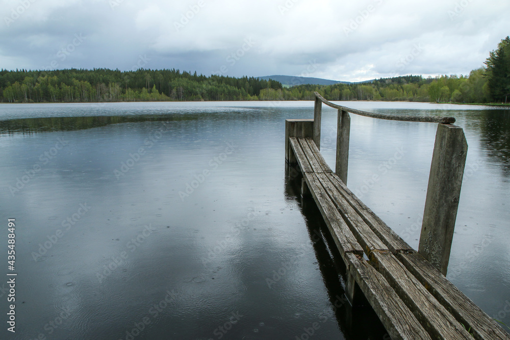 The old wooden pier over the surface of the pond in Czech nature during the cloudy day with a fine rain. 