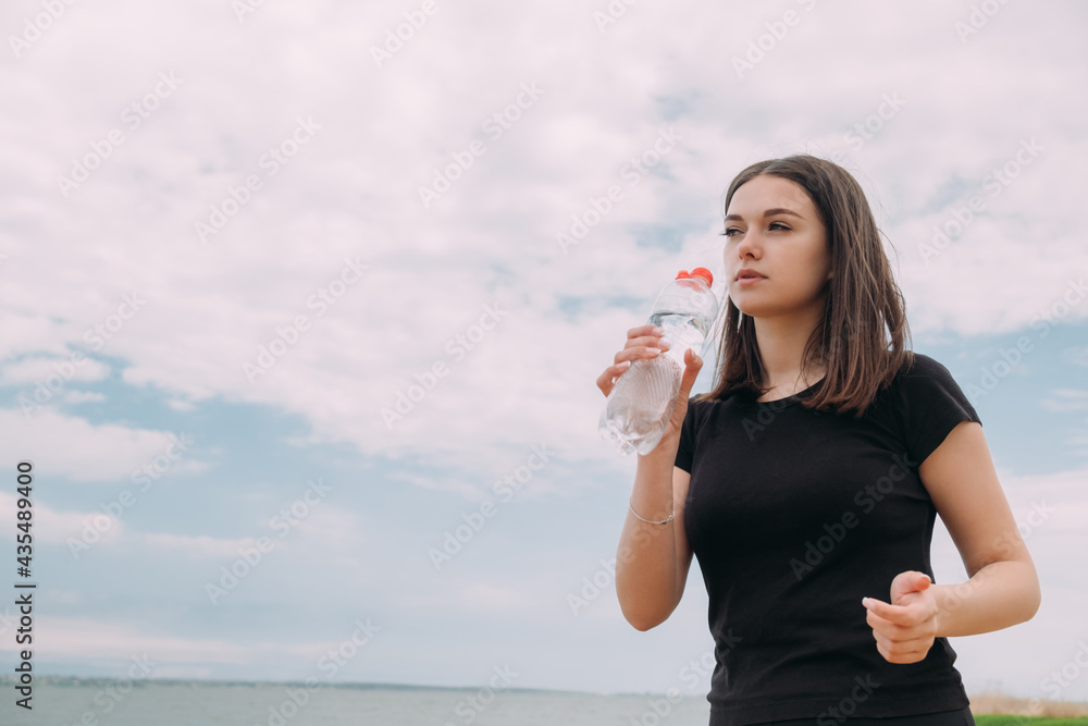A young girl of Caucasian appearance drinks water against the sky after jogging. A young girl of Caucasian appearance drinks water against the sky after jogging.