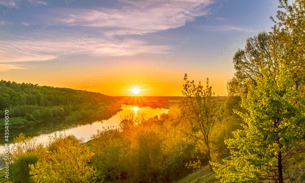 Scenic view at beautiful spring sunset on a shiny river valley with green branches, trees, bushes, grass, golden sun, calm water , blue cloudy sky and forest on a background, spring landscape