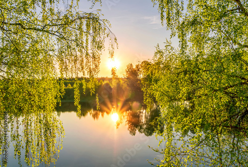 Scenic view at beautiful spring sunset on a shiny lake with green branches, birch trees, bushes, grass, golden sun rays, calm water ,deep blue cloudy sky and forest on a background, spring landscape
