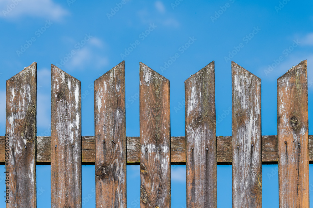 Top of old weathered pointed wood fence on a blue sky background. Vintage fencing detail. Shabby wooden planks with nature grains and knots, nails or pale smudges of white paint remainders. Side view. Top of old weathered pointed wood fence on a blue sky background. Vintage fencing detail. Shabby wooden planks with nature grains and knots, nails or pale smudges of white paint remainders. Side view.
