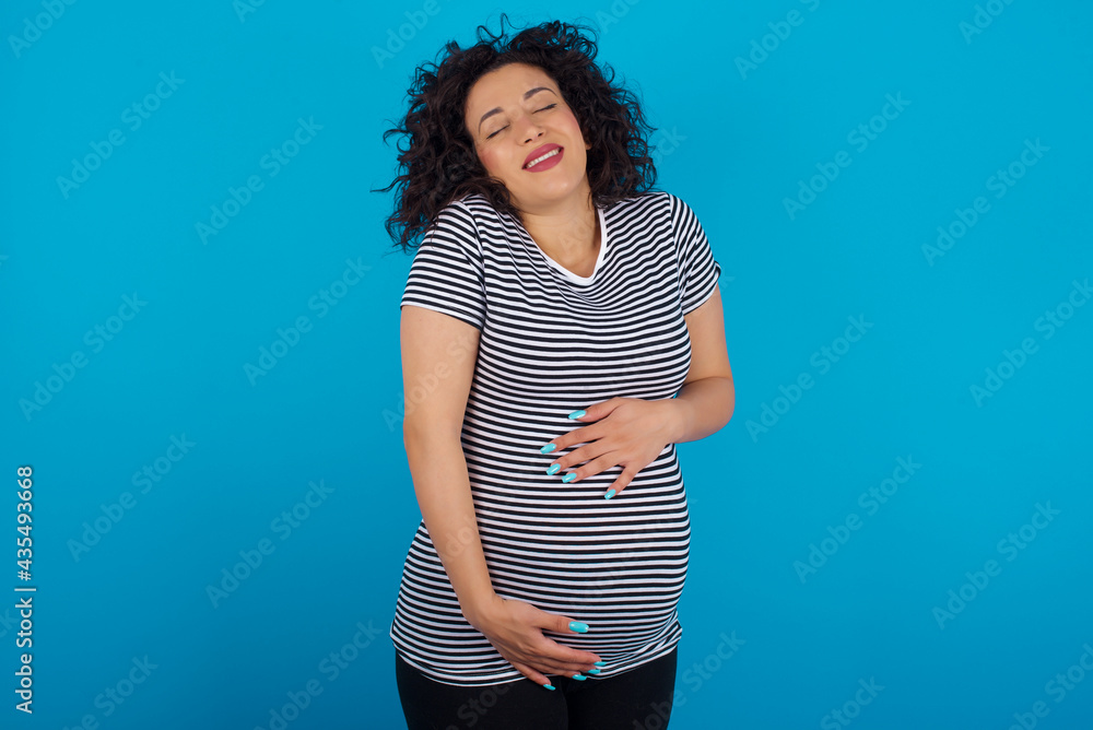 Positive young Arab pregnant woman wearing stripped T-shirt against blue wall with overjoyed expression closes eyes and laughs shows white perfect teeth Positive young Arab pregnant woman wearing stripped T-shirt against blue wall with overjoyed expression closes eyes and laughs shows white perfect teeth