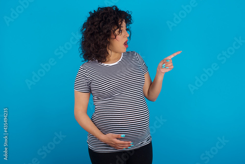 Stunned young Arab pregnant woman wearing stripped T-shirt  against blue wall with greatly surprised expression points away on copy space, indicates something