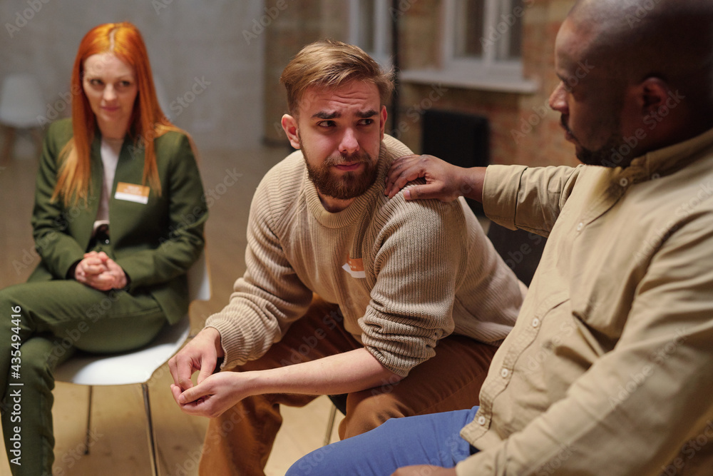 Obraz premium Young bearded man crying while looking at African guy supporting him at session