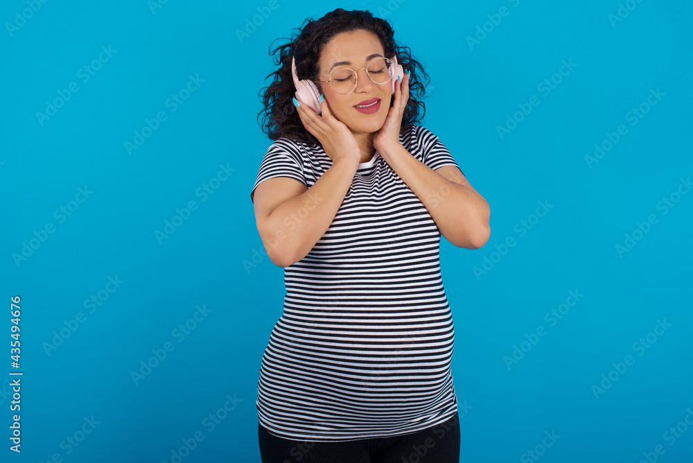 Pleased young Arab pregnant woman wearing stripped t-shirt standing against blue background enjoys listening pleasant melody keeps hands on stereo headphones closes eyes. Spending free time with music Pleased young Arab pregnant woman wearing stripped t-shirt standing against blue background enjoys listening pleasant melody keeps hands on stereo headphones closes eyes. Spending free time with music