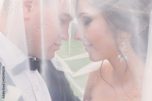 Stylish groom in a suit and a cute bride in a lace dress are standing under a long veil on their face and hugging close-up. Wedding portrait of smiling newlyweds.