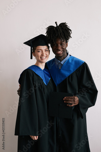 Vertical waist up portrait of young couple wearing graduation robes and smiling at camera