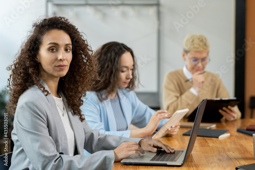 Attractive businesswoman with laptop sitting by table among co-workers