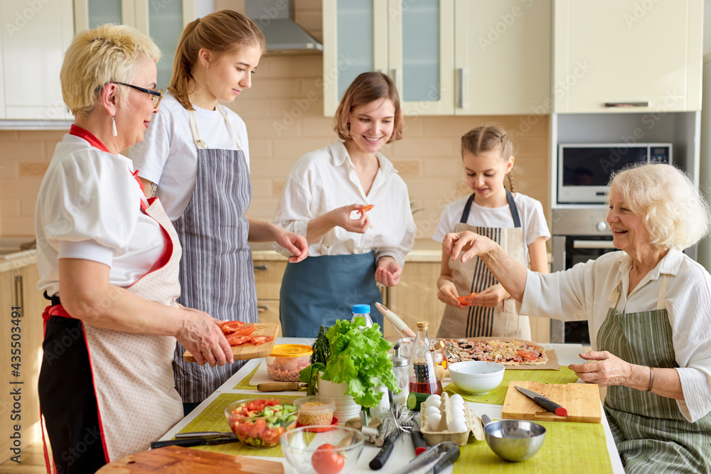 Caucasian family preparing delicious tasty pizza in bright modern kitchen, cutting products and adding ingredients, wearing apron.culinary concept. Grandmothers teach young girl to cook