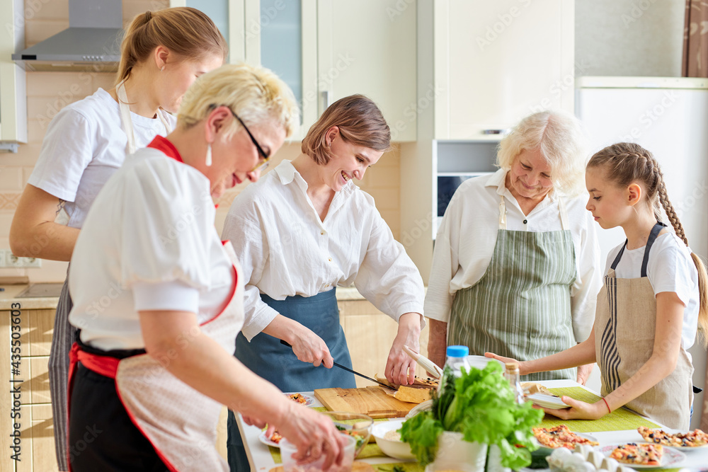 Side View Portrait of family preparing delicious tasty pizza in bright modern kitchen, cutting products and adding ingredients, wearing apron.culinary concept. Grandmothers teach young girl to cook Side View Portrait of family preparing delicious tasty pizza in bright modern kitchen, cutting products and adding ingredients, wearing apron.culinary concept. Grandmothers teach young girl to cook