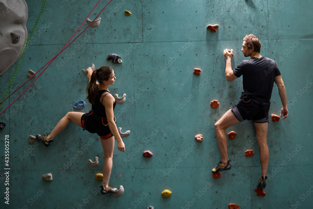 Athlete sporty woman and man in sportive outfit practicing rock climbing on artificial rock in sport club. Extreme sports and bouldering concept. - obrazy, fototapety, plakaty 