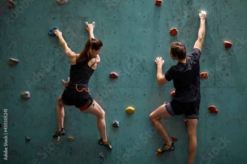 Professional woman and man mountaineers climbing artificial rock wall with belay at bouldering gym, in sportswear, rear view on professional sport people workout training photo