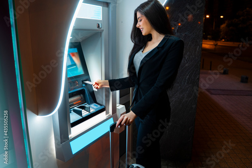 Obraz na plátně Pretty dark hair Caucasian woman with suitcase using ATM at night