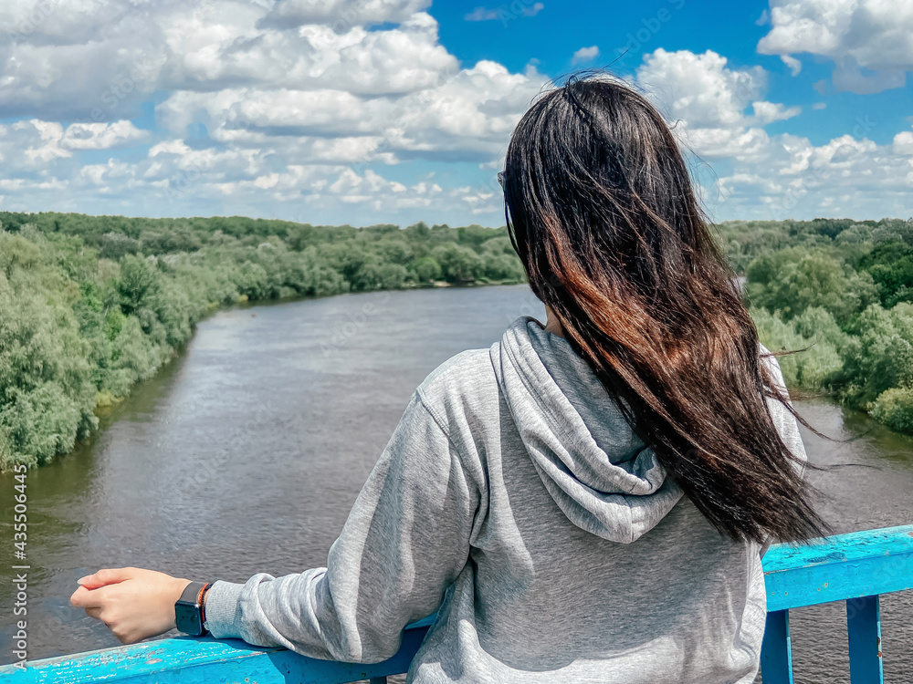 Fototapeta premium A dark-haired сaucasian girl stands with her back in a light gray jacket and looks towards the river and the forest in the city of Chernigov in Ukraine.
