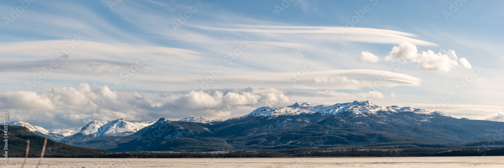Panorama of snow capped mountains in distance over a frozen lake and amazing, wispy clouds during spring time in northern Canada. 