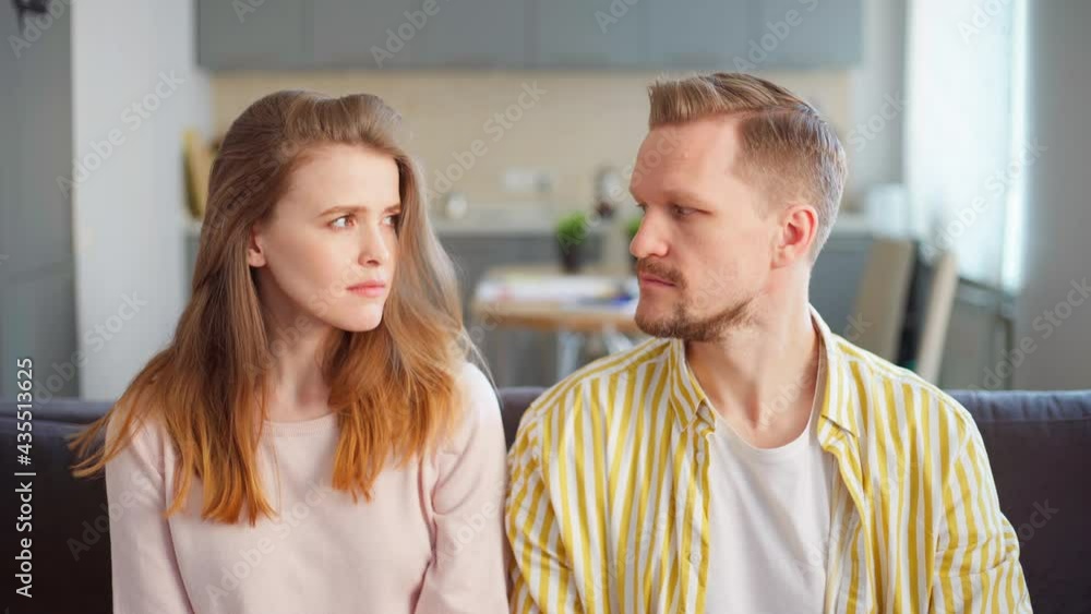 Unhappy sick and tired of relationship problems young couple sitting on sofa due to stay at home order and looking at camera. Man and woman in conflict looking at each other, female sighing sadly Unhappy sick and tired of relationship problems young couple sitting on sofa due to stay at home order and looking at camera. Man and woman in conflict looking at each other, female sighing sadly