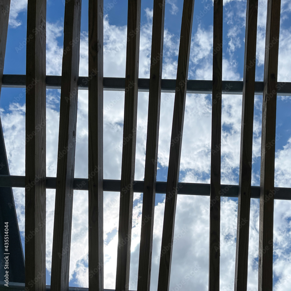 A wooden pergola roof at a park with bright blue skies and white clouds. A wooden pergola roof at a park with bright blue skies and white clouds.