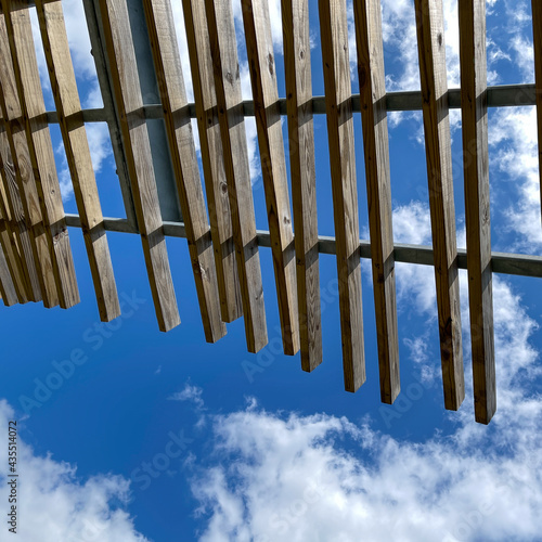 A wooden pergola roof at a park with bright blue skies and white  clouds.