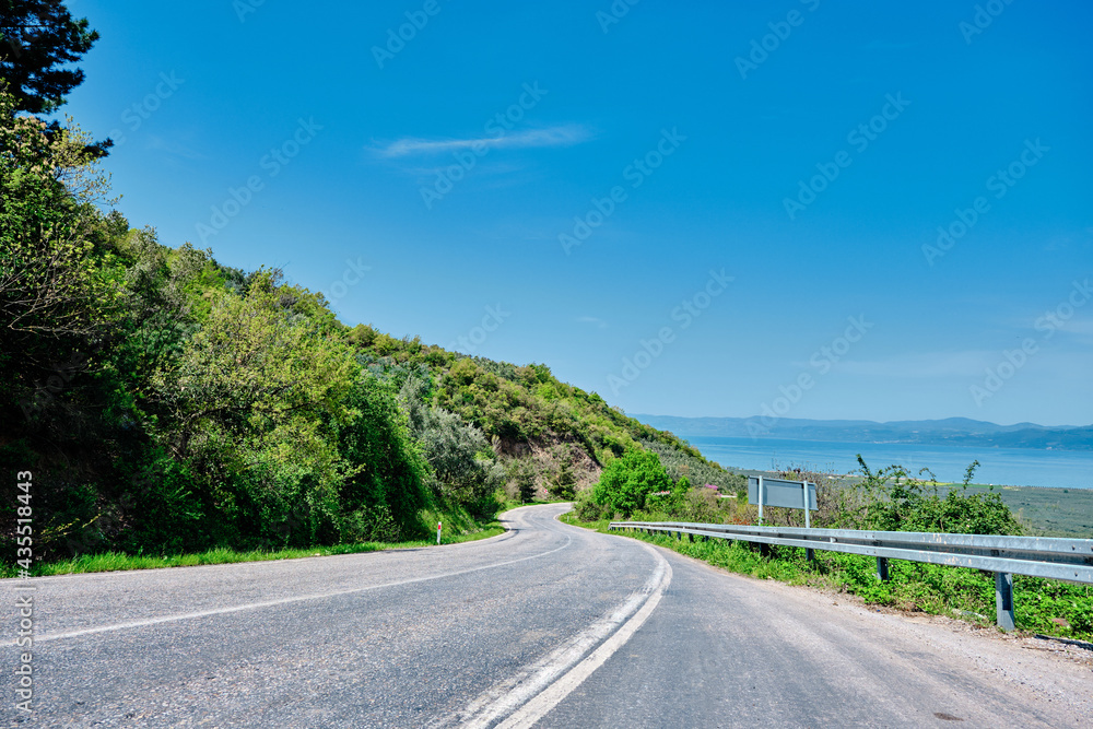 Low angle photo of asphalt road Bursa to Nicaea (iznik) during sunny day and road signs and protection fences next to road with small hills that is covered by many green plants 