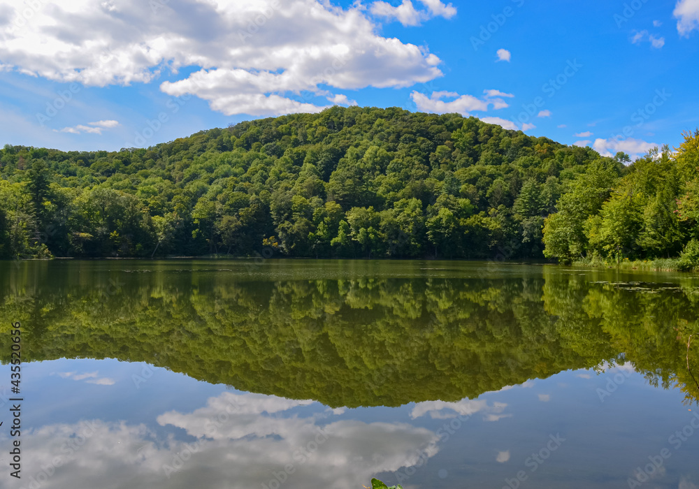 A lake in Vermont reflecting a wooded hillside and a blue summer sky with puffy clouds.  Copy space.