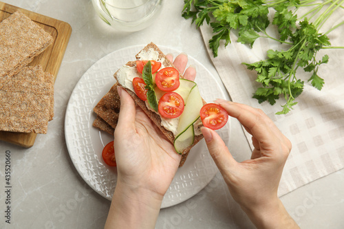 Woman eating fresh rye crispbread with cream cheese and vegetables at light grey marble table, top view photo