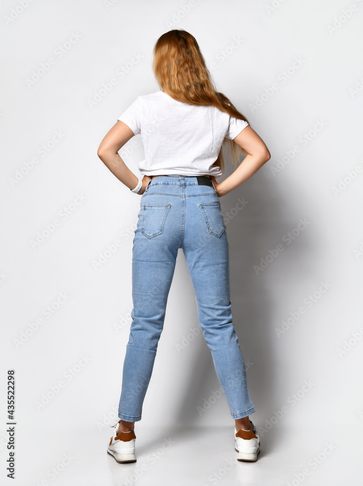 young skinny woman dressed in stylish blue jeans and white sneakers. Girl poses on a white background showing off pants. Concept of a new collection of denim clothing. Profile view. young skinny woman dressed in stylish blue jeans and white sneakers. Girl poses on a white background showing off pants. Concept of a new collection of denim clothing. Profile view.