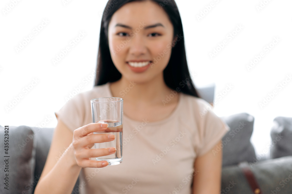 Close-up of defocused healthy happy pretty young asian woman sitting at home on sofa in casual clothes, leading a healthy lifestyle, holding a glass of water, looking at the camera, smiling Close-up of defocused healthy happy pretty young asian woman sitting at home on sofa in casual clothes, leading a healthy lifestyle, holding a glass of water, looking at the camera, smiling