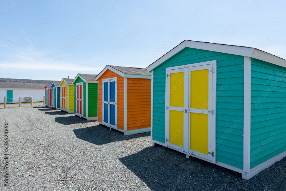 Multiple wooden sheds with colorful double doors. The buildings are blue, yellow and green storage buildings. The colorful huts have white trim on the edges. The background is a blue sky with clouds. 