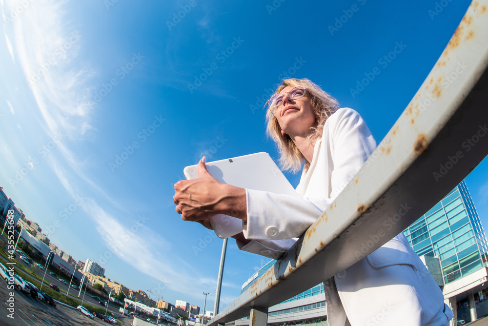 a blonde woman in a business white suit and round glasses stands with a white tablet in her hands against the background of the sky. horizontal photo. Half-length portrait. fish eye