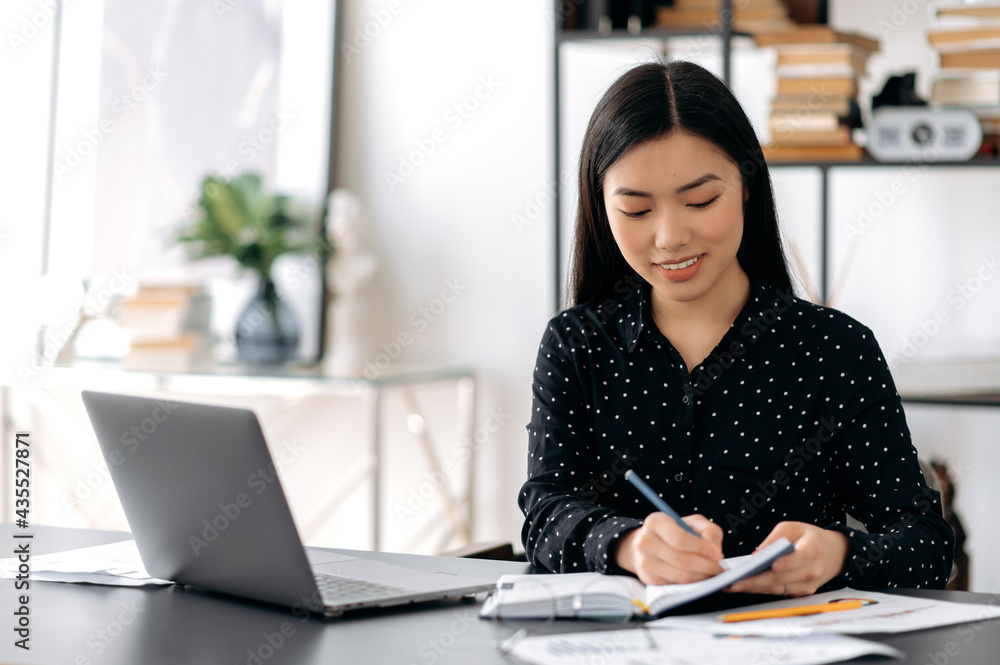 Online work with a laptop or distance learning. Young adult pretty female asian freelancer, manager or student, sitting at work desk, using laptop for work or study, taking notes during online webinar Online work with a laptop or distance learning. Young adult pretty female asian freelancer, manager or student, sitting at work desk, using laptop for work or study, taking notes during online webinar