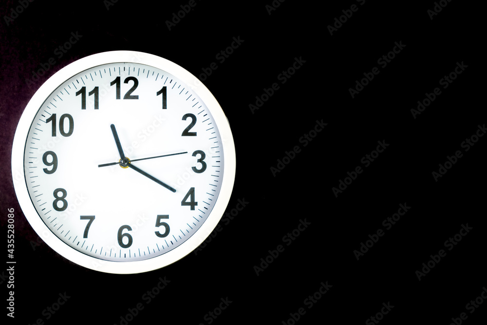Close-up of a modern fashioned white Wall Clock with numerals on the clock face. Clock with white frame on a black background. Pointers showing the time. front view of the Watch out of center Close-up of a modern fashioned white Wall Clock with numerals on the clock face. Clock with white frame on a black background. Pointers showing the time. front view of the Watch out of center